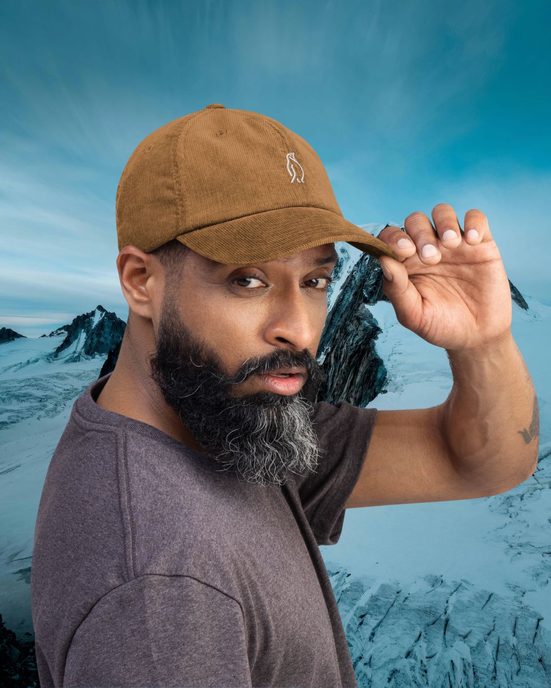 Cord Cap Driftwood mit weißer Pinguin-Stickerei – Mann hält  Cap Schirm vor verschneiter Bergkulisse